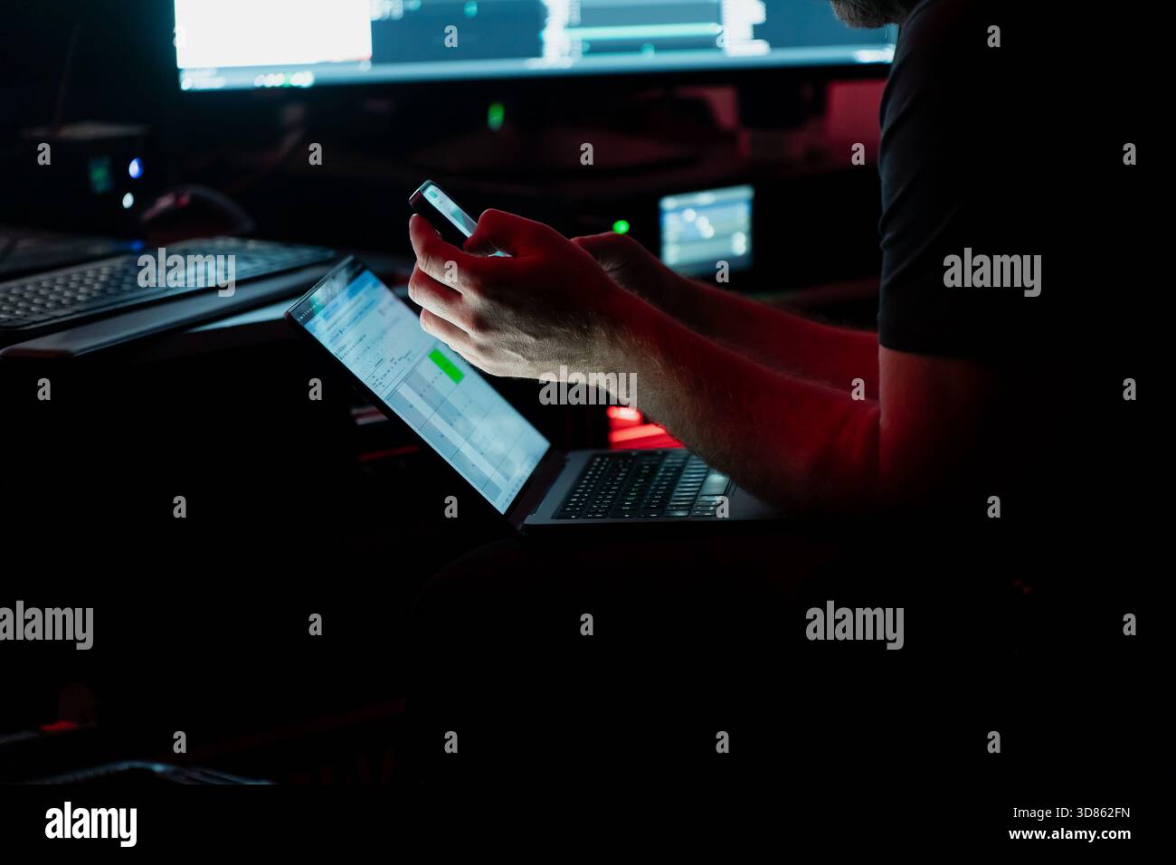 Bühnentechniker, der digitale Tools auf Laptop und Smartphone bei schlechten Lichtverhältnissen in Backstage-Steuerungsumgebungen während des technischen Workflows verwaltet Stockfoto
