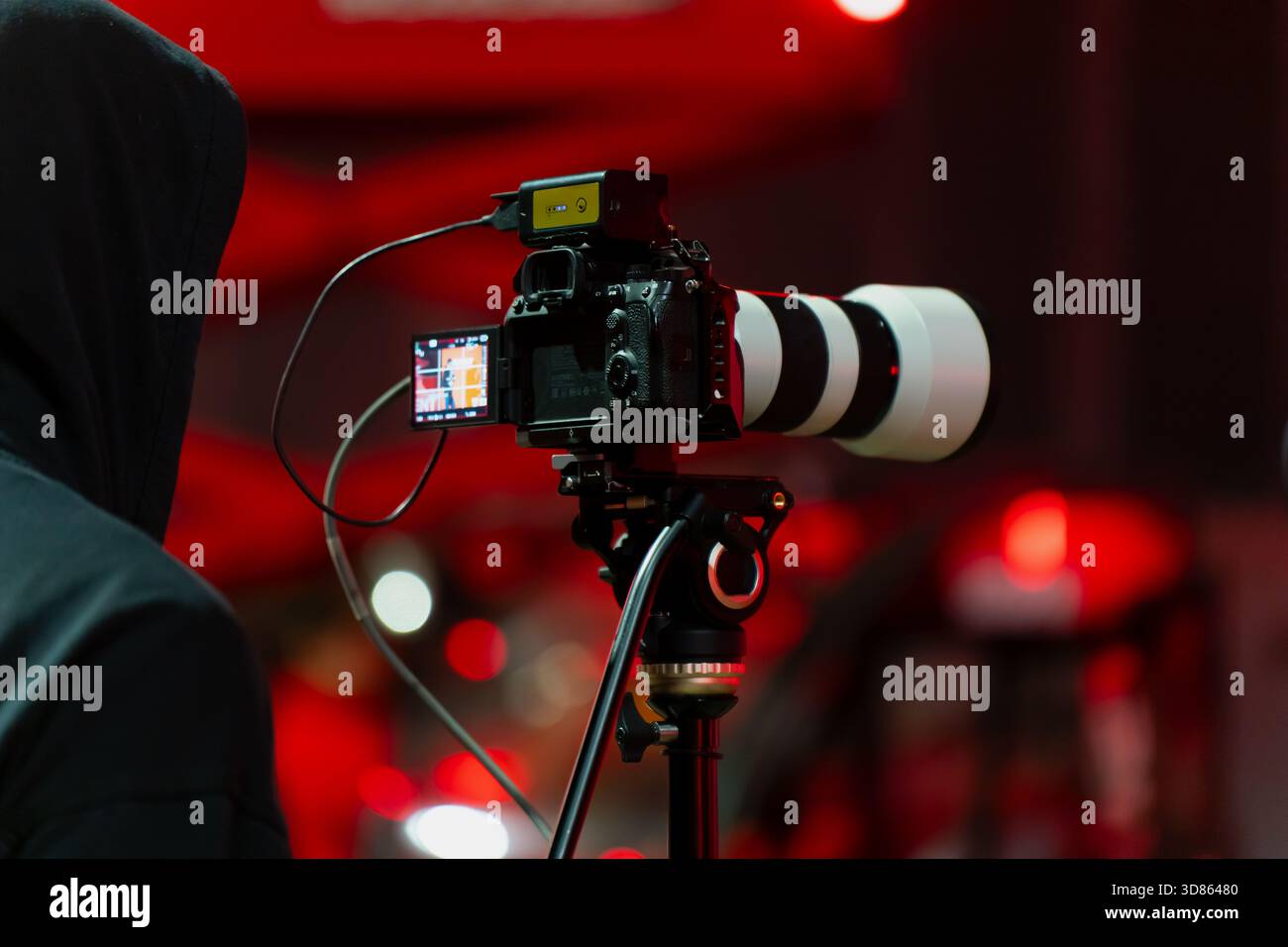Filmeinrichtung mit einer professionellen Kamera auf einem Stativ und einem Bediener, der Inhalte in einer rot beleuchteten Umgebung, die typisch für Event-Marketing ist, live aufzeichnet Stockfoto