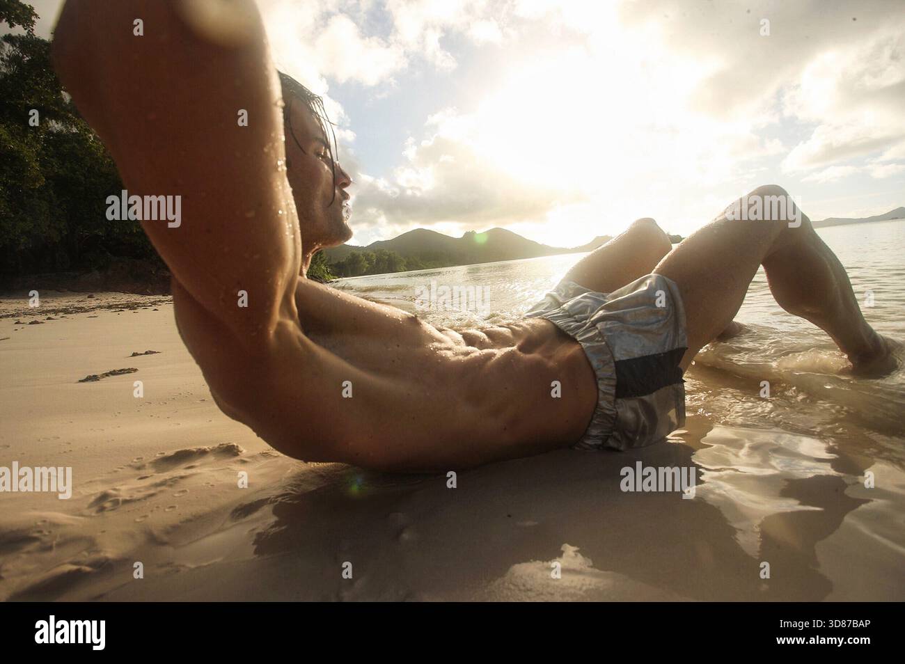 Nahaufnahme eines Mannes, der während eines goldenen Sonnenuntergangs am Strand ein abs-Workout macht. Das männliche sportliche Model macht Sit-ups, die Szene zeigt Bewegung. Stockfoto