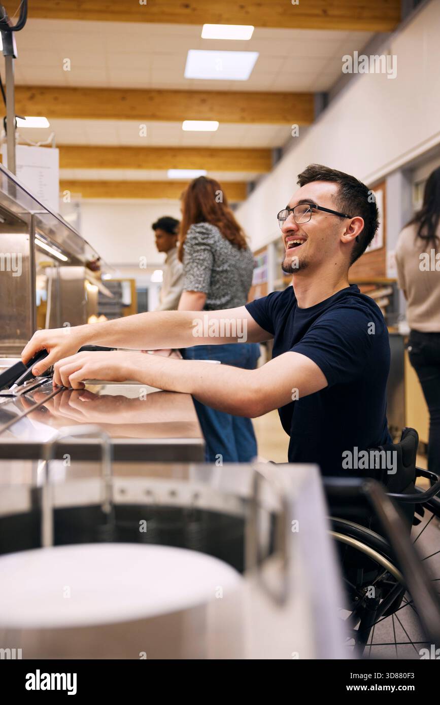 Glücklicher junger Mann, der Essen aus dem Container nimmt, während er auf Rollstuhl in der Schulcafeteria sitzt Stockfoto