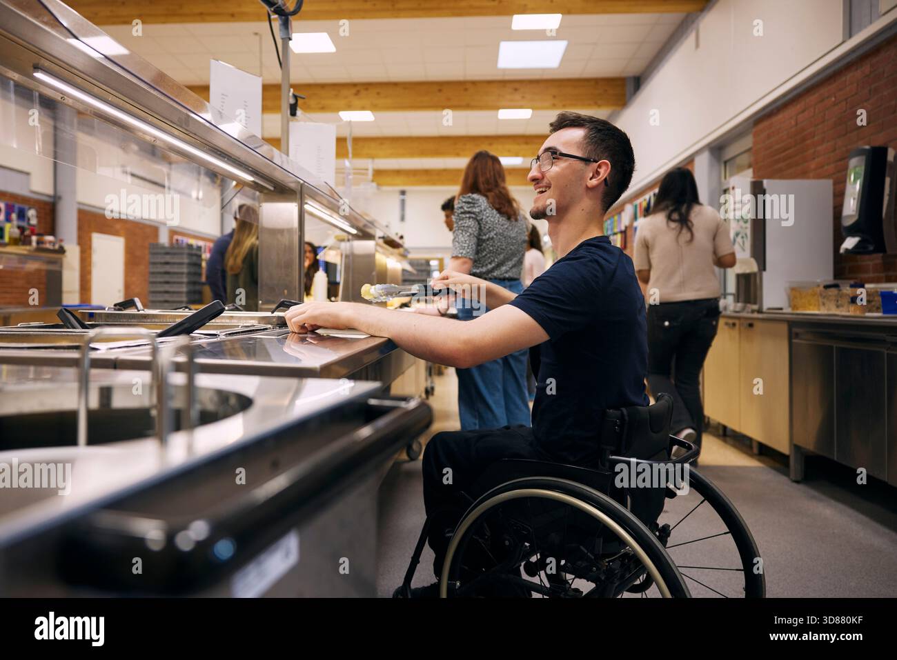 Glücklicher junger Mann, der im Rollstuhl sitzt und Essen aus dem Container nimmt, während des Mittagessens in der Schulcafeteria Stockfoto