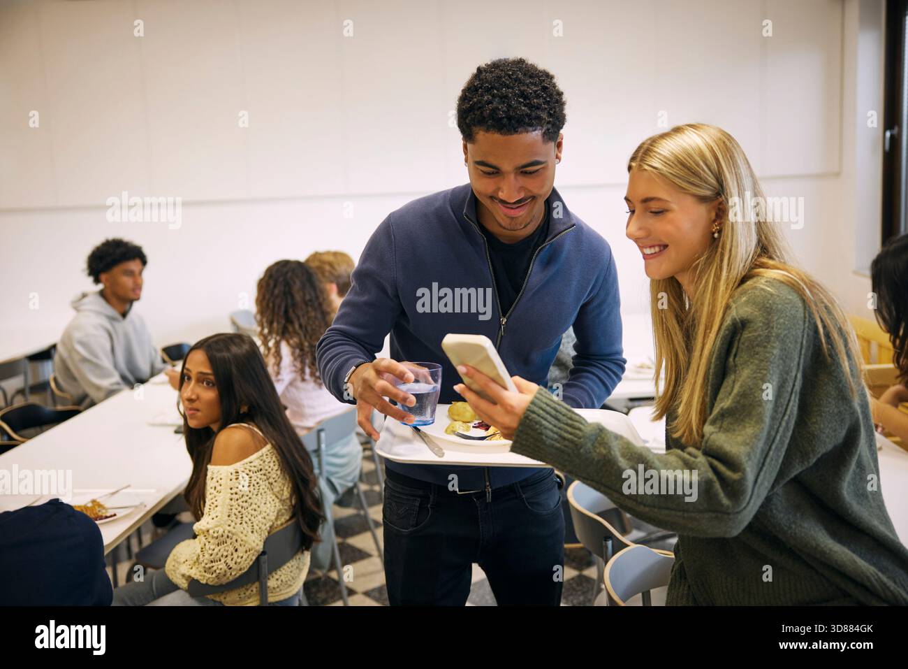 Lächelndes blondes Mädchen, das einem männlichen Freund das Smartphone zeigt, während der Mittagspause im High School Café mit Essensteller steht Stockfoto