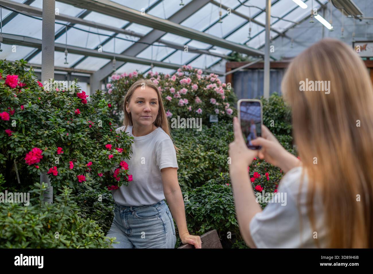 Die junge Frau steht inmitten farbenfroher Blumen in einem Gewächshaus, während ihre Tochter im Teenageralter ein Foto macht, das ihre fröhliche Beziehung und ihre gemeinsame Ex hervorhebt Stockfoto