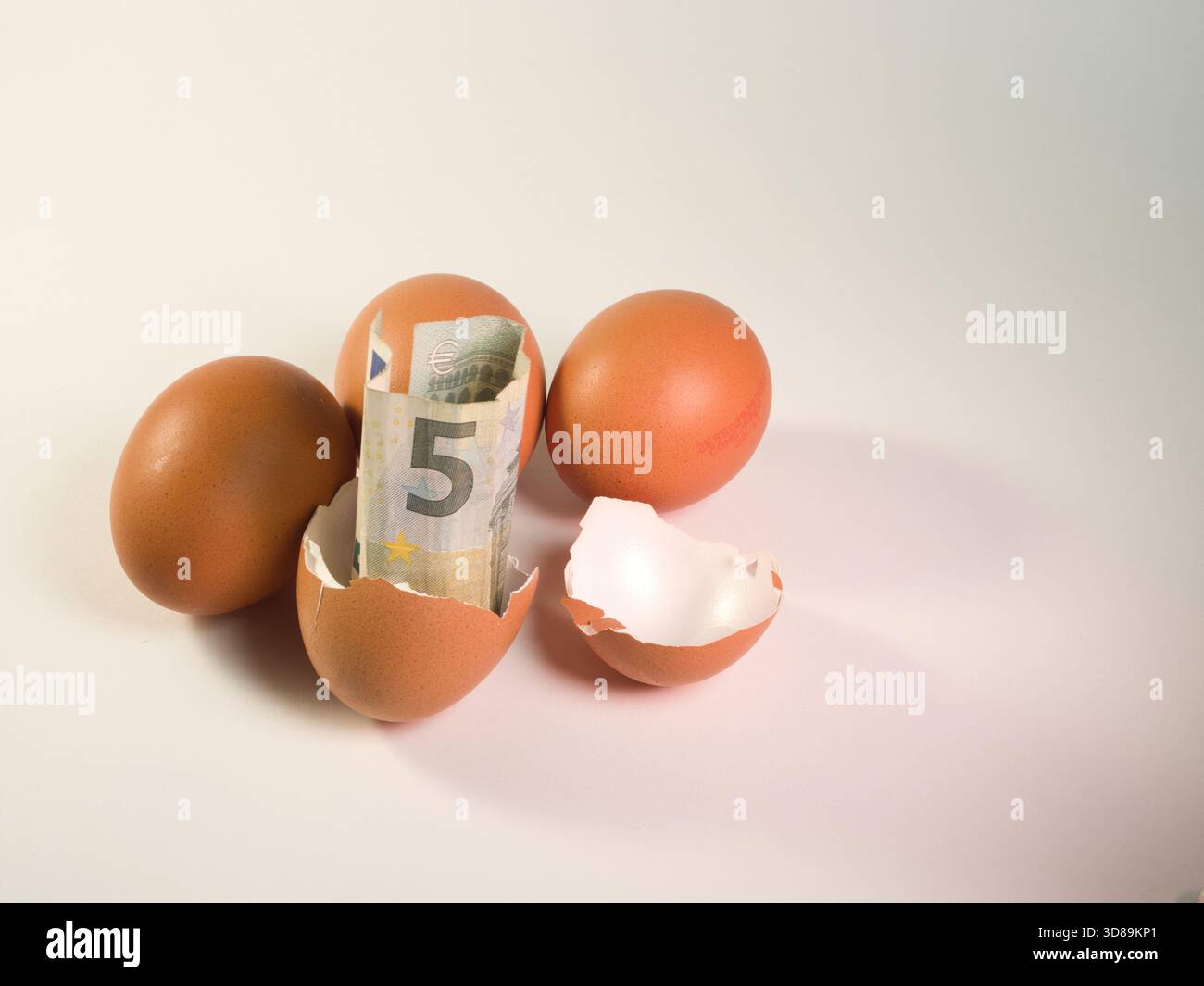 Eurogeld in Broken Eggshell - fünf Euro Schein und Münzen in einer kaputten Eierschale mit ganzen Eiern auf weiß. Symbolisiert Finanzen, unerwartete Kosten oder ich Stockfoto