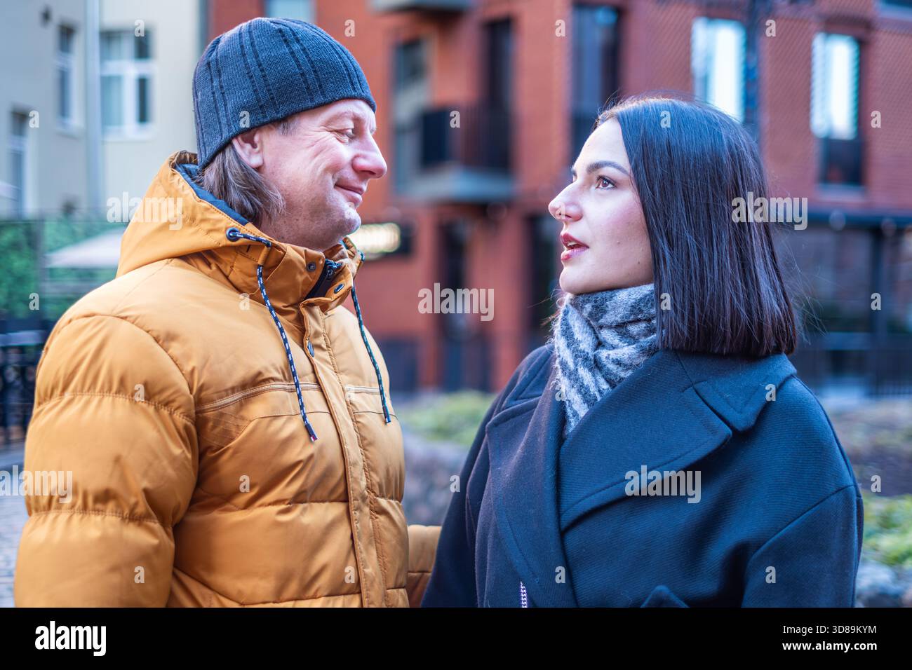 Mann und Frau unterhalten sich im Freien, tragen Winterjacken, umgeben von einer einladenden urbanen Umgebung, die ein warmes und intimes Ambiente schafft Stockfoto