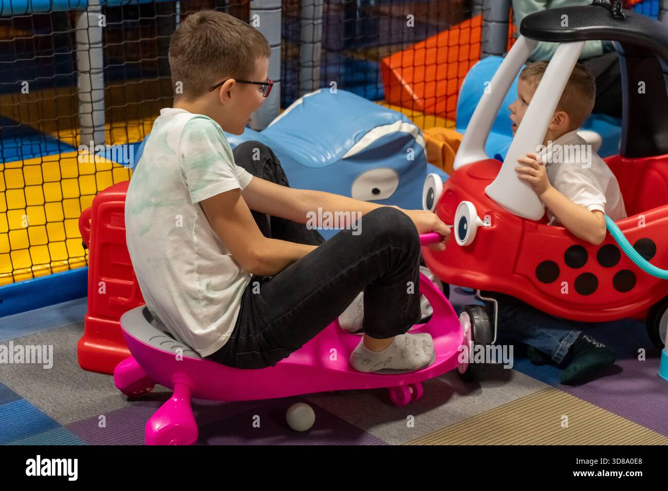 Zwei Brüder, die auf dem Spielplatz mit bunten Spielzeugautos fahren, mit einem älteren Jungen auf einem Wackelwagen und einem jüngeren in einem gemütlichen Coupé. Geschwister Spielspaß, Indoor-Eingang Stockfoto
