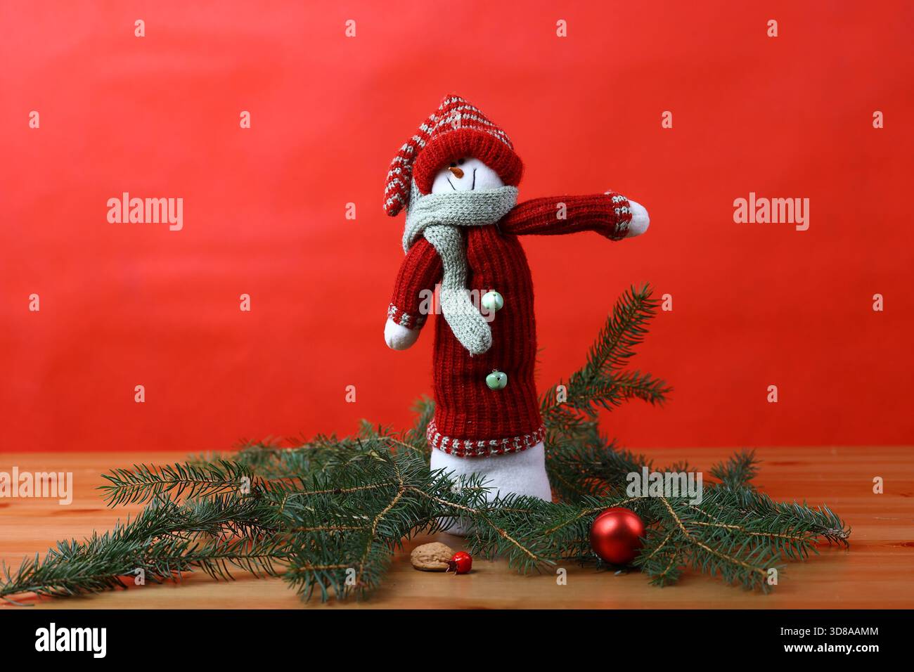 Ein weicher, gestrickter Schneemann mit rotem Outfit und Schal, umgeben von frischen Fichtenzweigen mit weichen Weihnachtsschmuck. Stockfoto