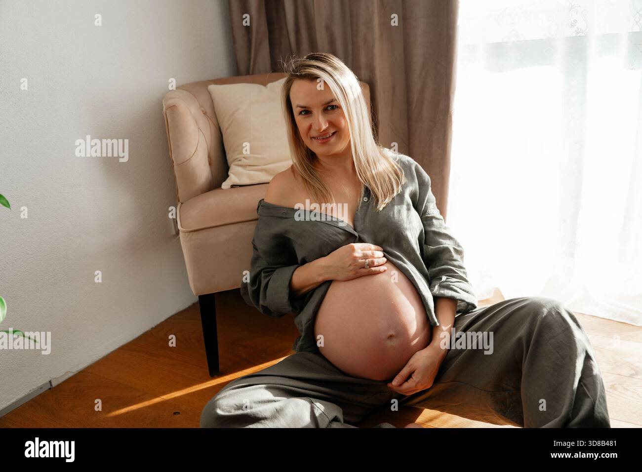 34 Wochen, Ein sanfter Mutterschaftsmoment mit einer schwangeren Frau, die bei hellem Tageslicht auf dem Boden in der Nähe eines Sofas sitzt. Ruhige Töne und eine entspannte Haltung sorgen für einen Stockfoto