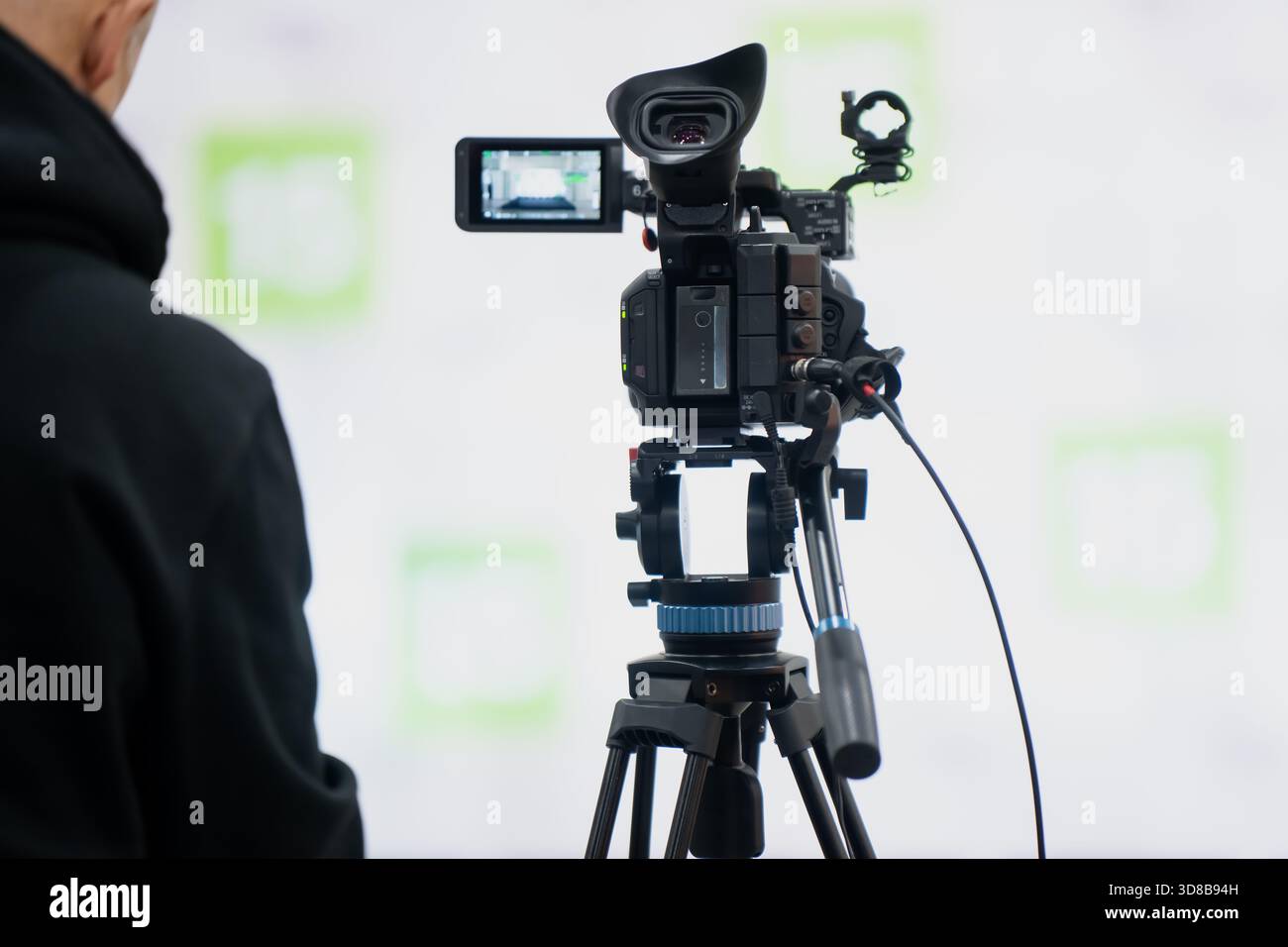 Filmeinrichtung mit einer professionellen Videokamera auf einem Stativ, das vor eine helle Kulisse gerichtet ist, während ein Bediener in der Nähe in einer Innenproduktion steht Stockfoto