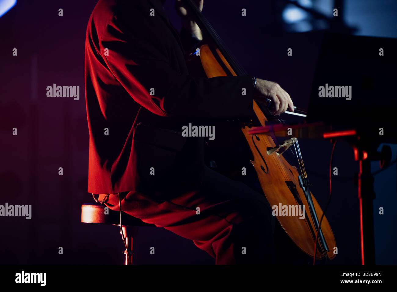 Nahaufnahme eines Musikers, der auf einem Cello spielt, während er auf einer Bühne sitzt, die mit lebhaftem Licht beleuchtet ist und von einem Laptop begleitet wird, der live genutzt wird Stockfoto