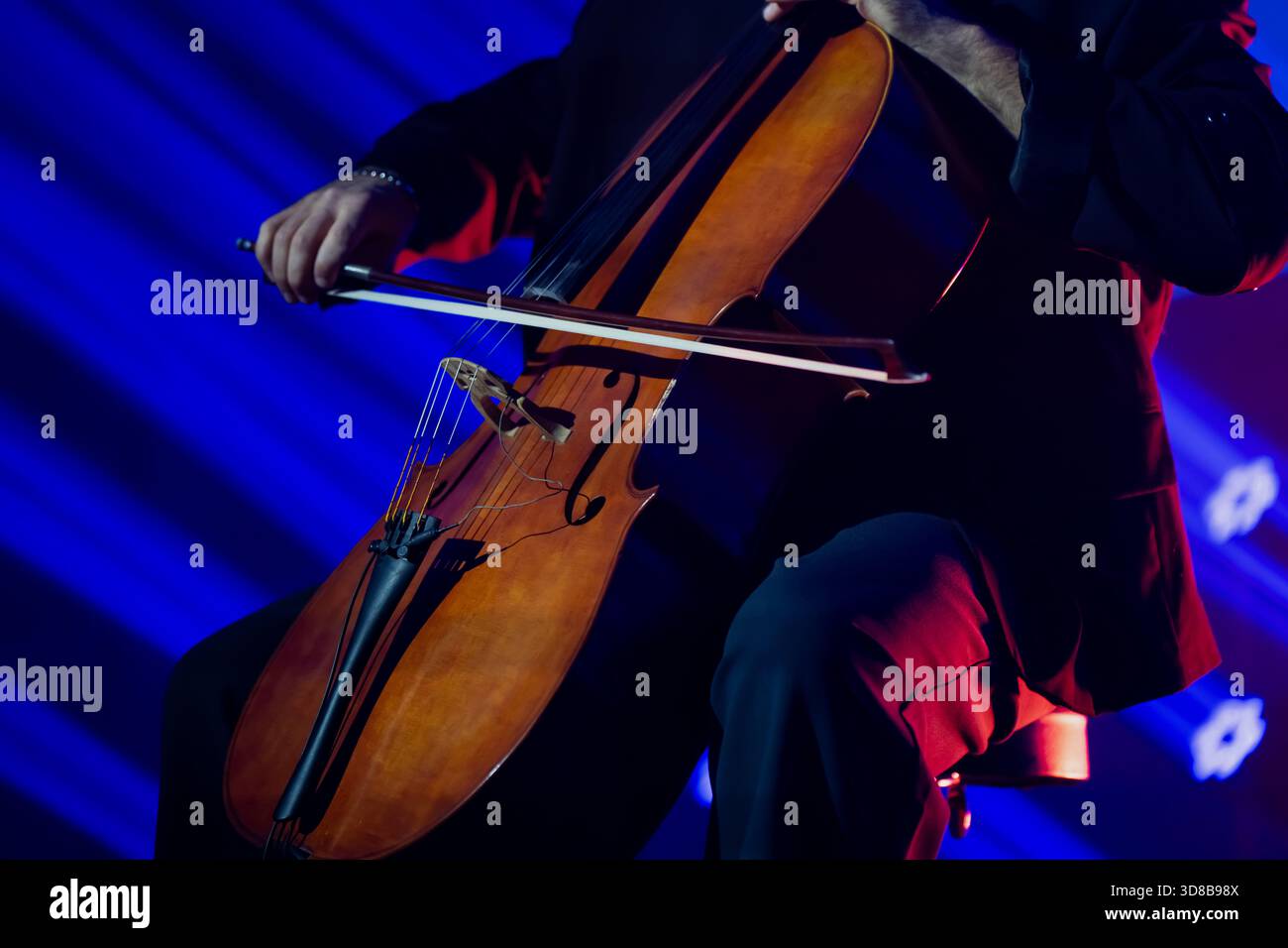 Musiker, der auf einem Cello unter dramatischer blauer und roter Bühnenbeleuchtung auftritt, mit Nahaufnahme des Fokus auf das Instrument und die Bogenbewegung in einer Live-Konzertumgebung Stockfoto