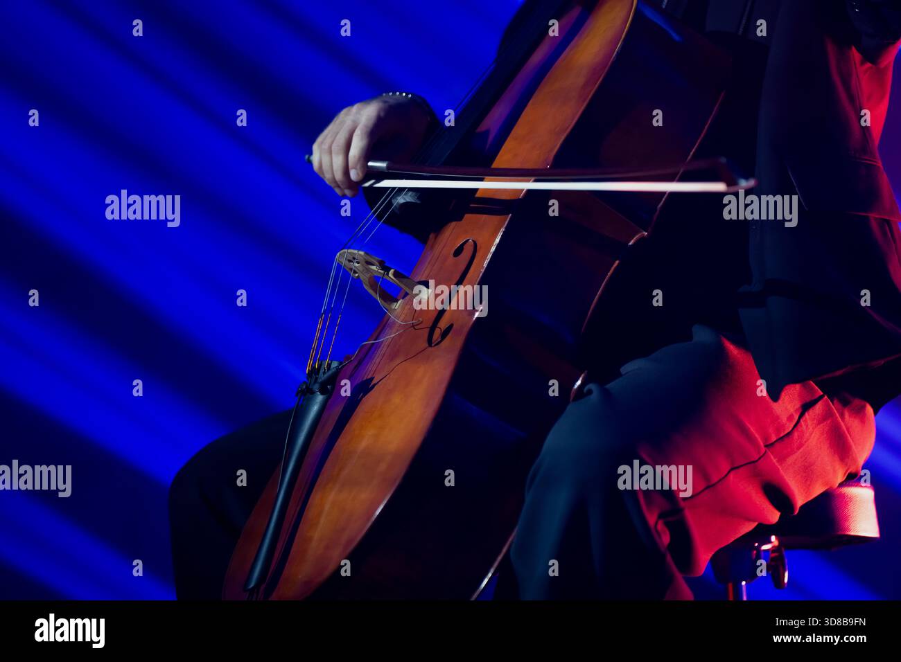 Live-Bühnenszene mit einem Cellisten in einem Anzug, der mit einem polierten hölzernen Cello gegen dynamische blaue Lichtstrahlen auftritt und ein lebendiges Konzert-atmos erzeugt Stockfoto