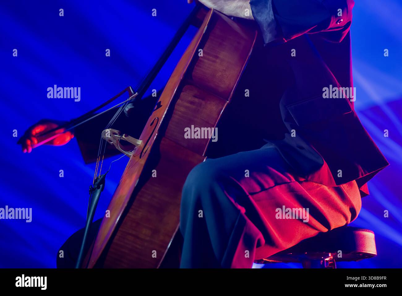 Cellist tritt auf der Bühne mit starken blauen Lichtstrahlen auf, die das Instrument beleuchten und warme Highlights über den Anzug des Musikers werfen Stockfoto
