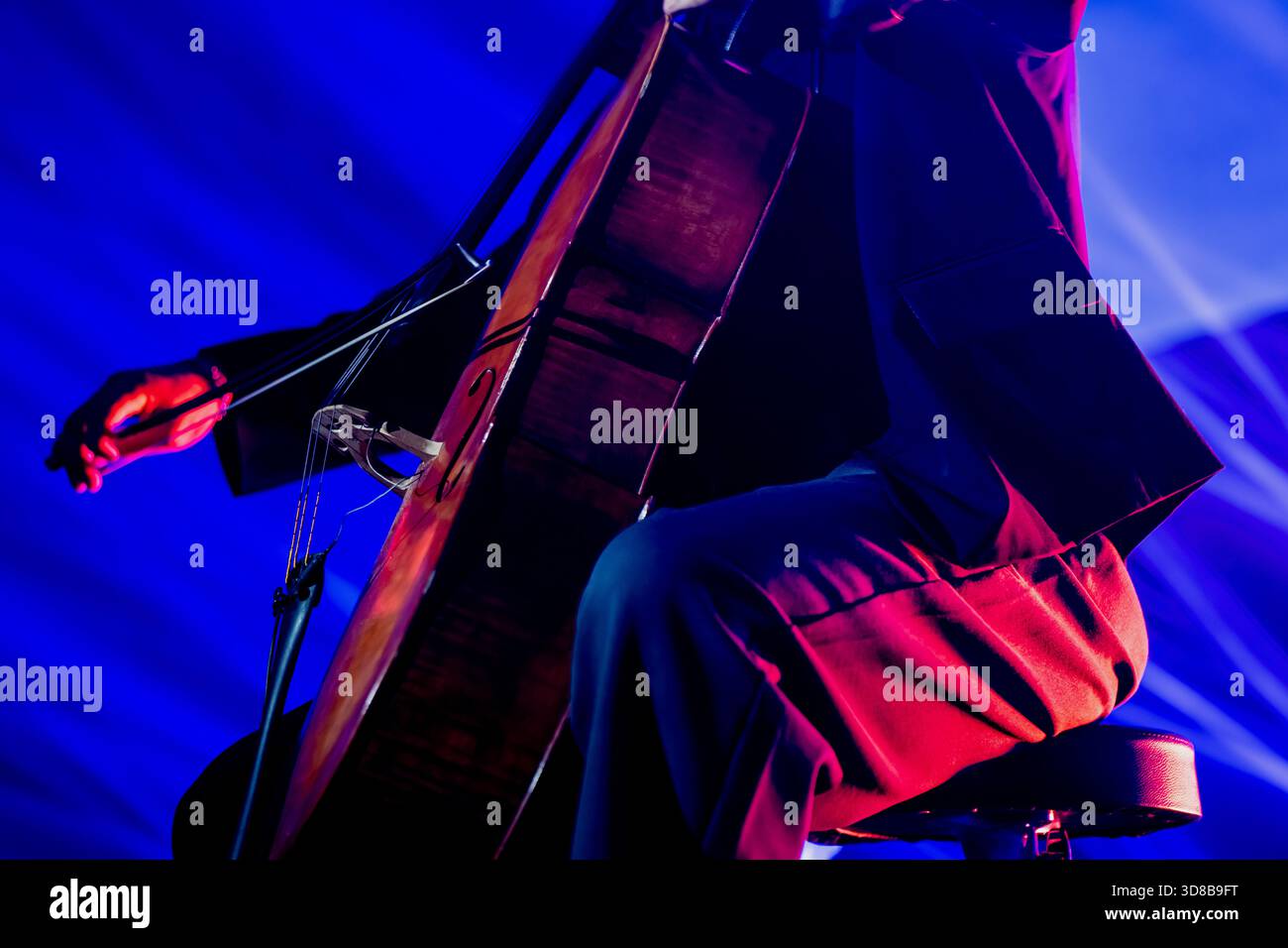Nahaufnahme eines Musikers, der ein poliertes hölzernes Cello unter dramatischer blauer Bühnenbeleuchtung mit sichtbarer Bogenbewegung und ausdrucksstarker Haltung während eines li spielt Stockfoto