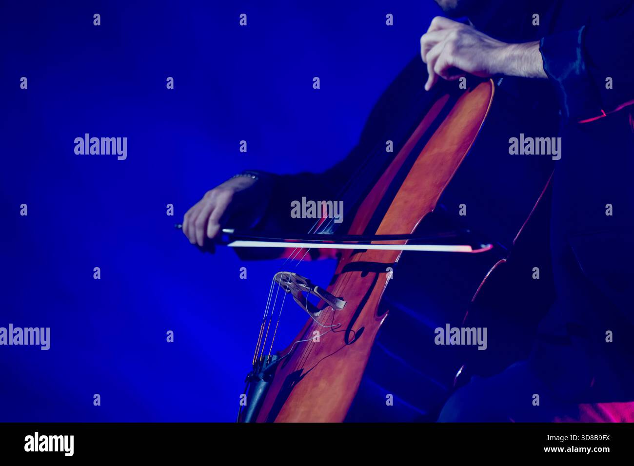 Nahaufnahme eines Cellospielers, der von intensiven blauen Lichtstrahlen beleuchtet wird, die die warmen Töne und die dynamische Bogenbewegung des Instruments hervorheben Stockfoto
