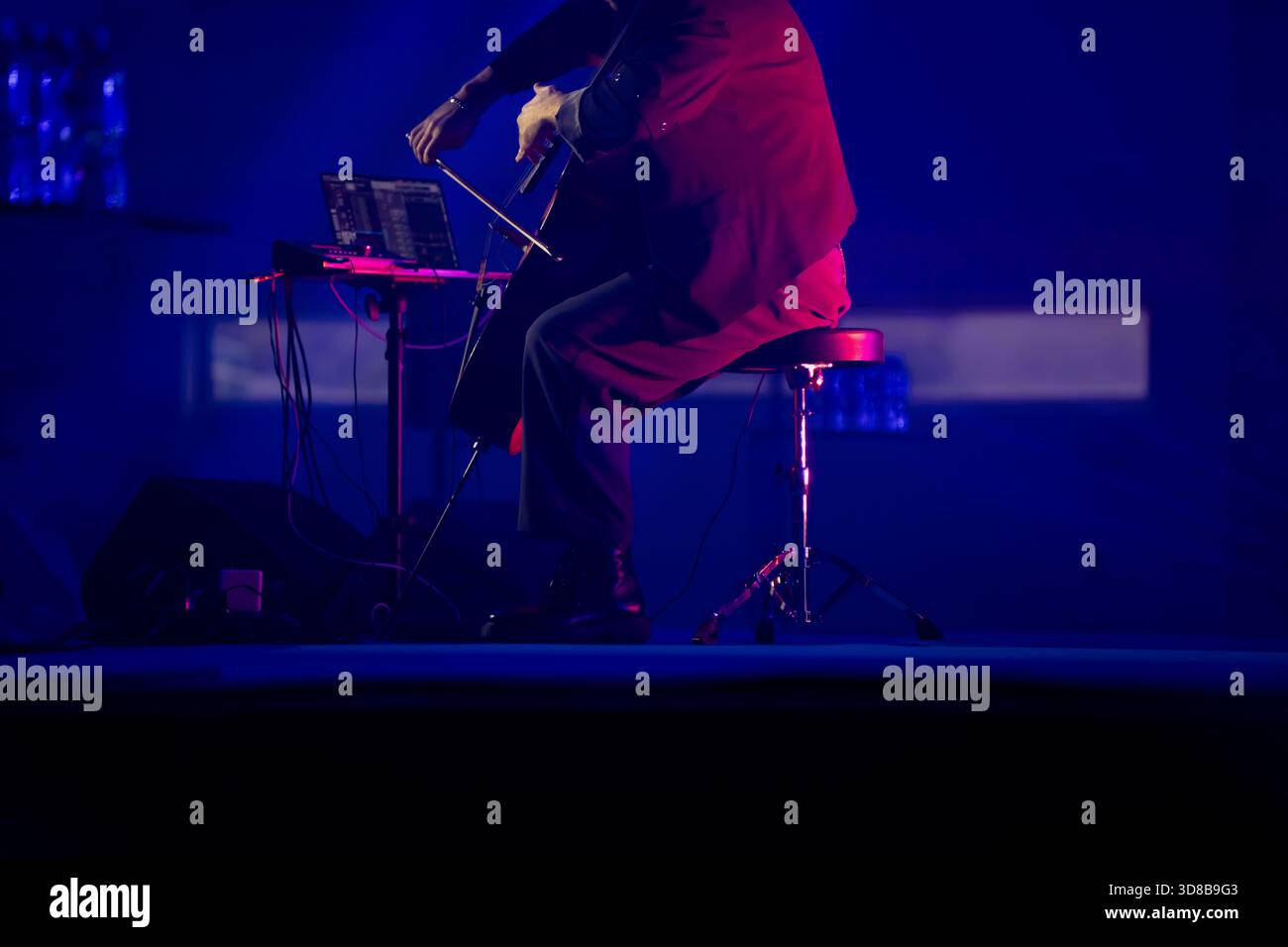 Sitzender Musiker auf der Bühne mit Cello und elektronischer Einrichtung einschließlich Laptop, Kabeln und Hocker, beleuchtet durch tiefblaues Licht während einer Li Stockfoto