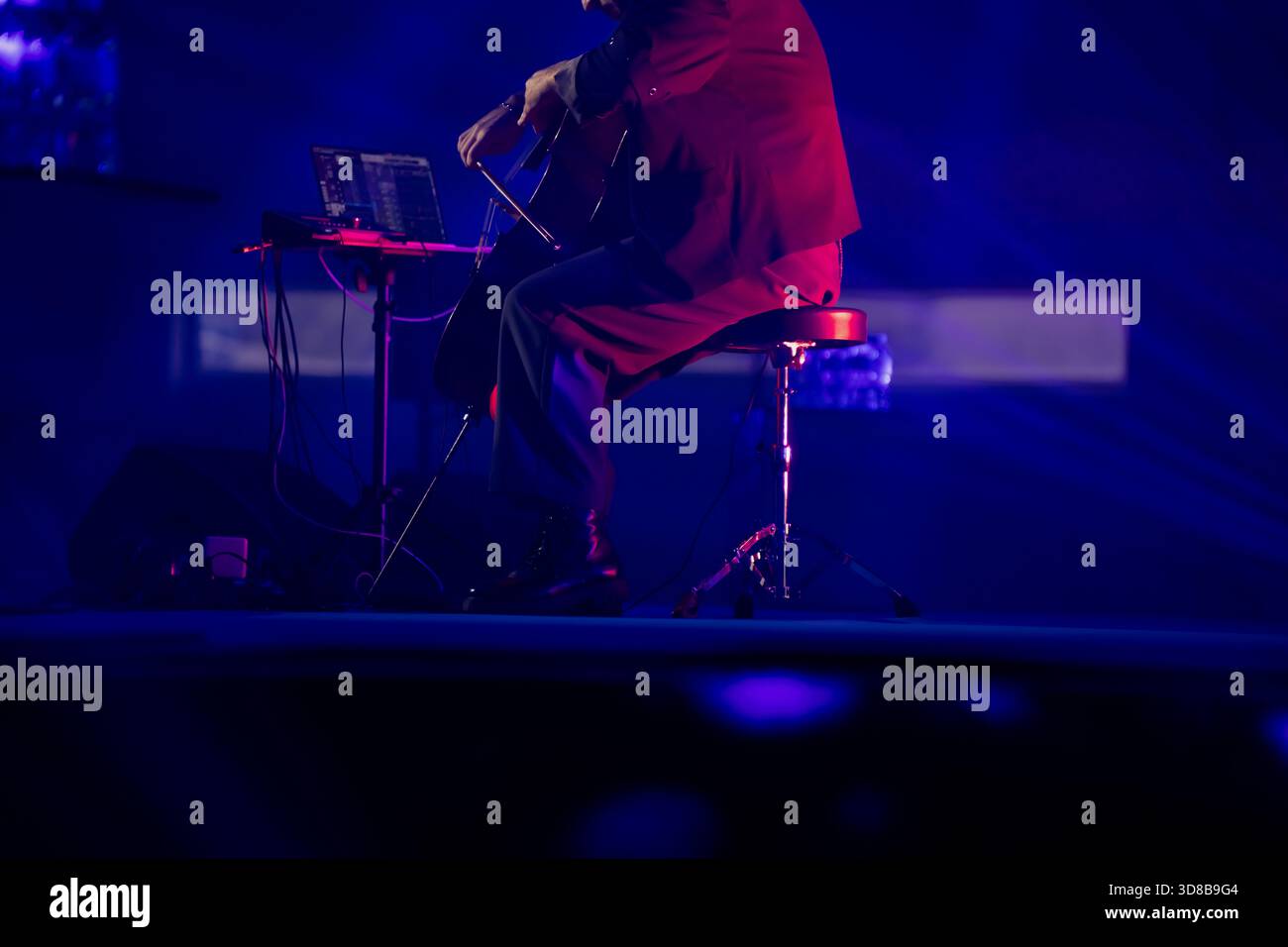 Live-Bühnenszene mit einem Cellisten, der mit einem Bogen und elektronischer Ausrüstung auf einem Performance-Hocker sitzt, unter dramatischer blauer Beleuchtung und Konzert-Atmosphäre Stockfoto