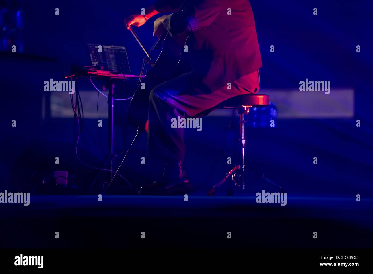 Musiker, der an einem elektronischen Arbeitsplatz mit Kabeln und Ausrüstung auf einer schwach beleuchteten Bühne mit starken Blautönen und fokussiertem per Cello spielt Stockfoto