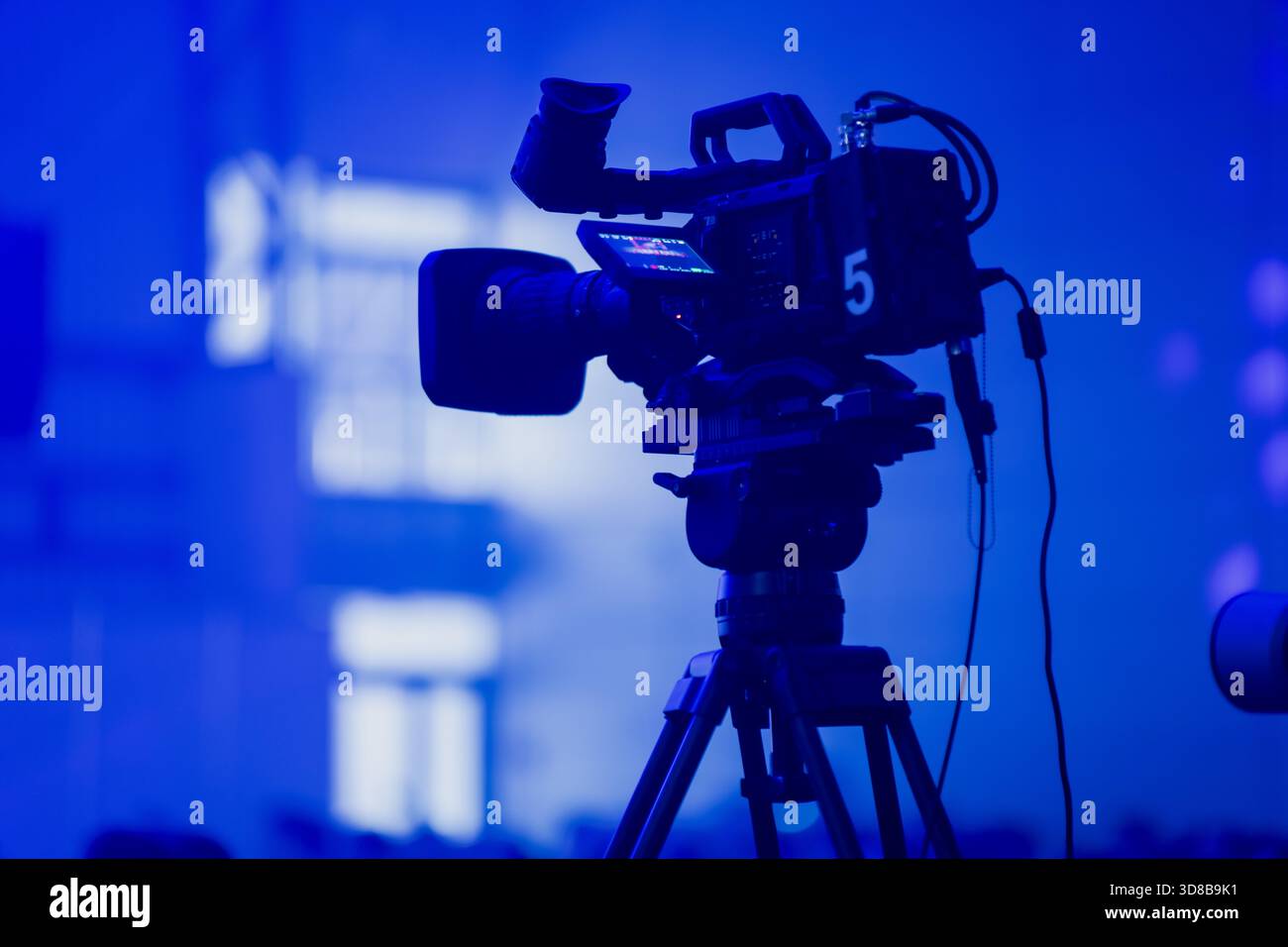 Professionelle Broadcast-Kamera auf einem Stativ, beleuchtet durch tiefblaue Eventbeleuchtung, die technische Ausrüstung für moderne Live-Übertragungen hervorhebt Stockfoto