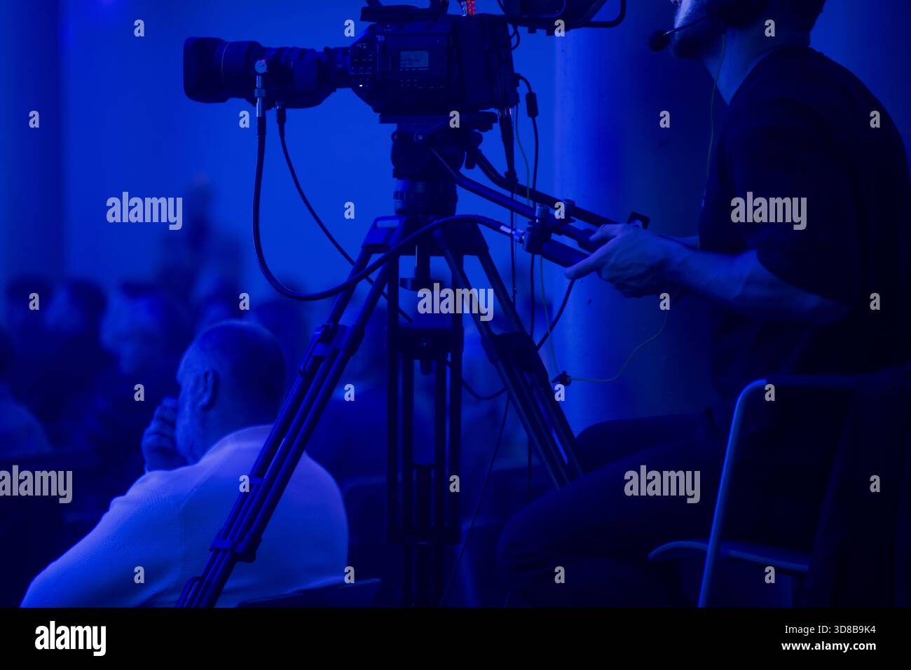 Der Kamerabediener filmt ein Ereignis von einer Stativplattform aus unter tiefblauer Beleuchtung mit dem Publikum im Hintergrund, um eine professionelle Veranstaltung zu veranschaulichen Stockfoto