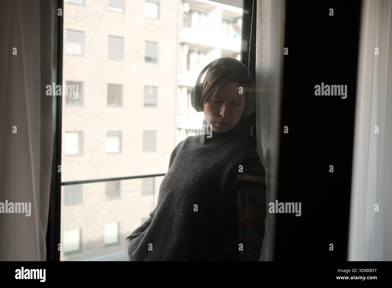 Eine Frau mit Kopfhörern lehnt sich mit sanfter Reflexion an ein Fenster und schafft eine nachdenkliche und introspektive Szene. Stockfoto