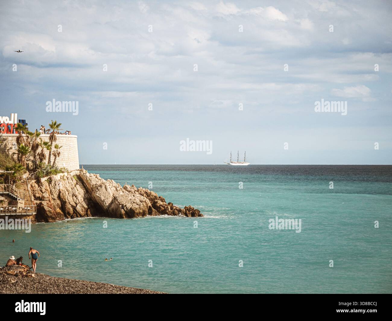 Felsige Küste von Nizza mit türkisfarbenem Wasser, Strandgängern, einem weit entfernten Großschiff und dem Schild „I Love Nice“ unter einem teilweise bewölkten Himmel. Stockfoto