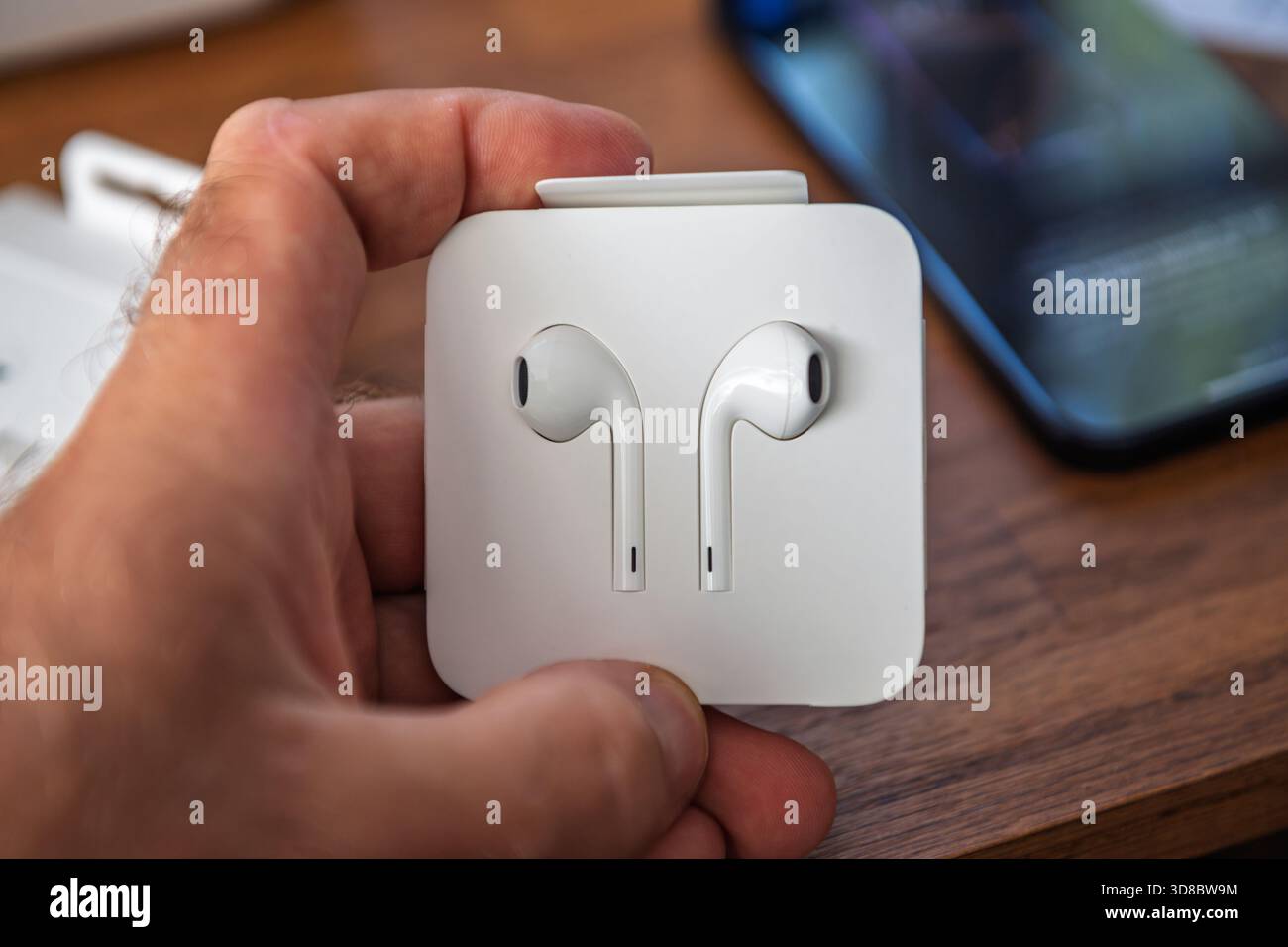Paris, Frankreich – 2. September 2025: Die neuen Apple EarPods mit USB-C-Anschluss auf der Verpackungskarte im Einzelhandel während des Auspackens in der Hand halten. Das iPhone ist im Hintergrund verschwommen und hebt das kabelgebundene Audiozubehör und das Produktdesign hervor. Stockfoto