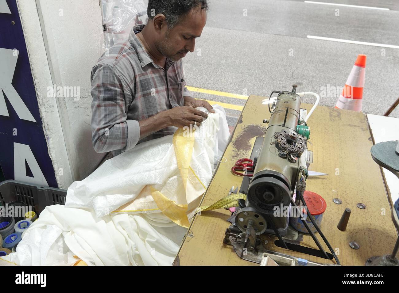 Little India, Singapur, 9. September 2025 männlicher Handwerker repariert ein weißes Kleidungsstück, während seine Nähmaschine in einer Kleiderwerkstatt stillsteht Stockfoto