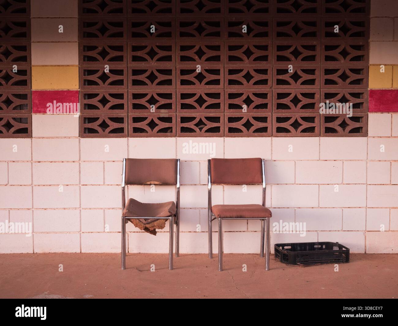 Zwei alte Stühle, einer kaputt, sitzen an einer weiß, rot und gelb gekachelten Wand Stockfoto