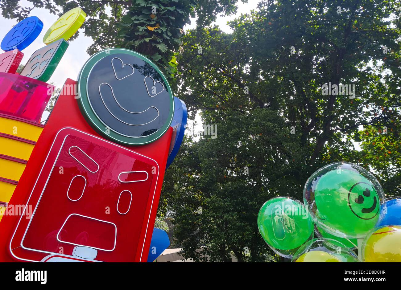Eine verspielte Außenschau mit einer riesigen Emoji-Skulptur und transparenten Ballons, die während eines fröhlichen öffentlichen Festes in einem belebten Park angeordnet werden Stockfoto