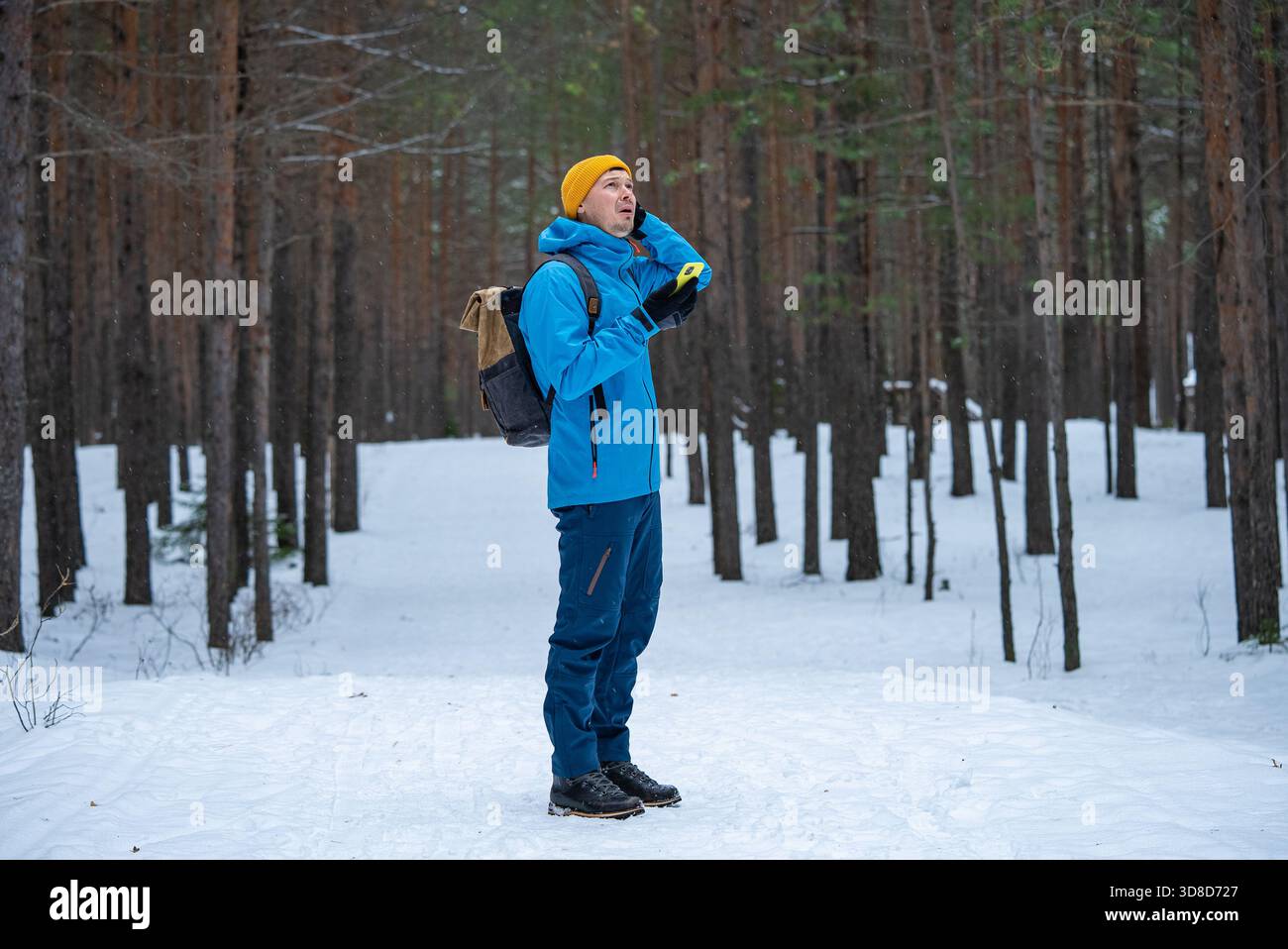 Mann in Winterkleidung und Rucksack, der besorgt aufblickt, während er ein Handy hält, kein Signal oder keine Verbindung hat und sich in einem verschneiten Kiefernwald verloren und desorientiert fühlt Stockfoto