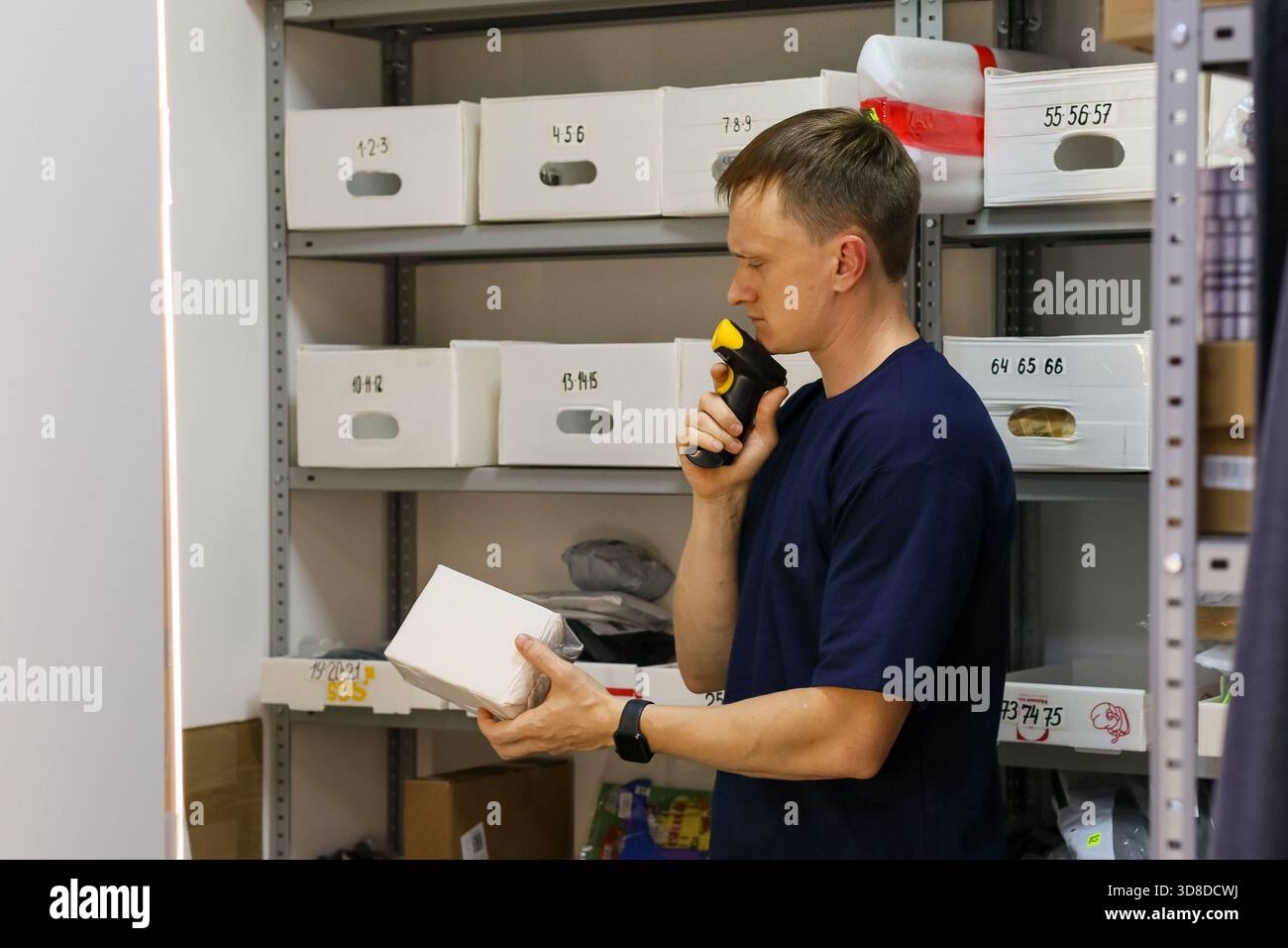 Mann in blauem T-Shirt scannt weiße Verpackung mit Gerät, während er neben Metallregalen steht. Stockfoto