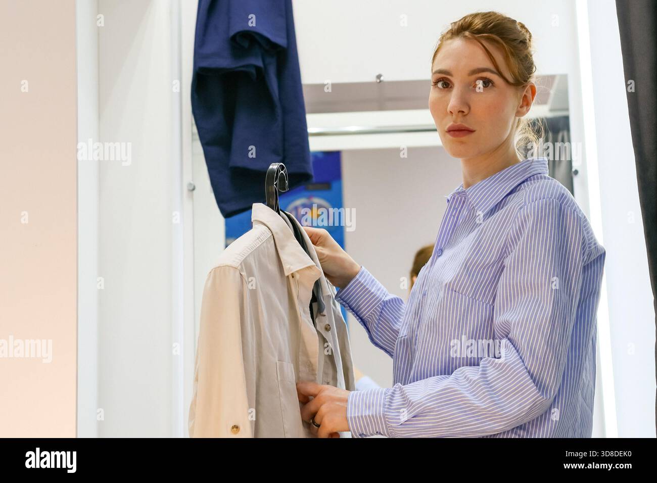 Junge Frau in gestreiftem Hemd, die auf eine helle Jacke auf einem Kleiderbügel blickt, dunkles Hemd im Hintergrund. Stockfoto
