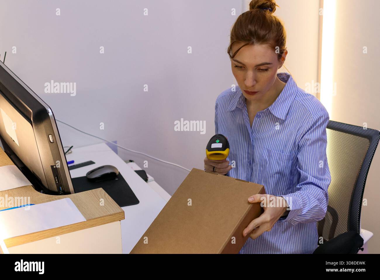 Die junge Frau verwendet einen Strichcode-Leser auf einem geneigten Karton am Schreibtisch mit Computer und Zubehör. Stockfoto