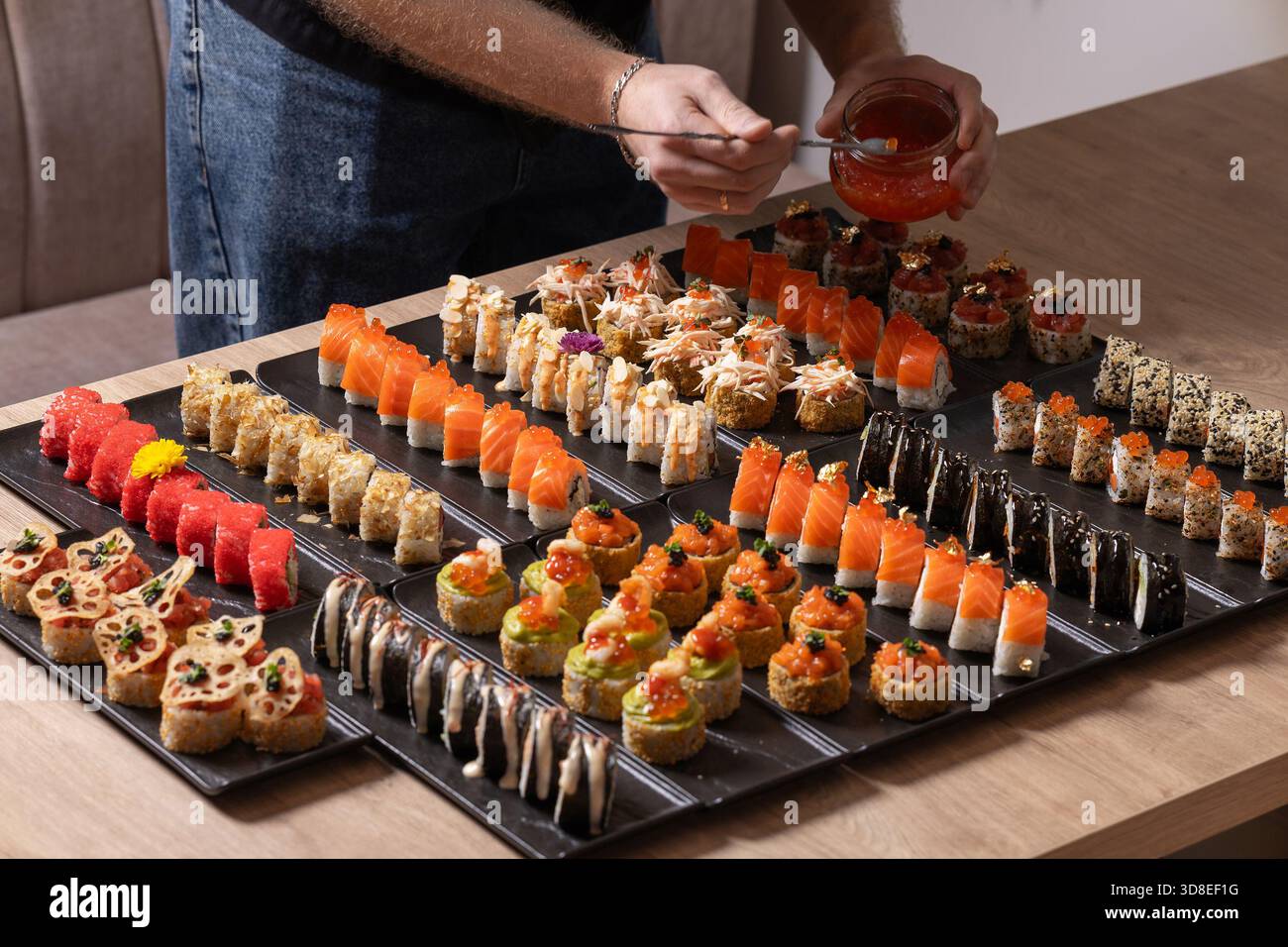 Eine Person spritzt elegant Sauce auf eine luxuriöse, große Gourmet-Platte mit verschiedenen Sushi-Brötchen, einschließlich Lachs, rotem Kaviar und essbarem Goldtoppin Stockfoto