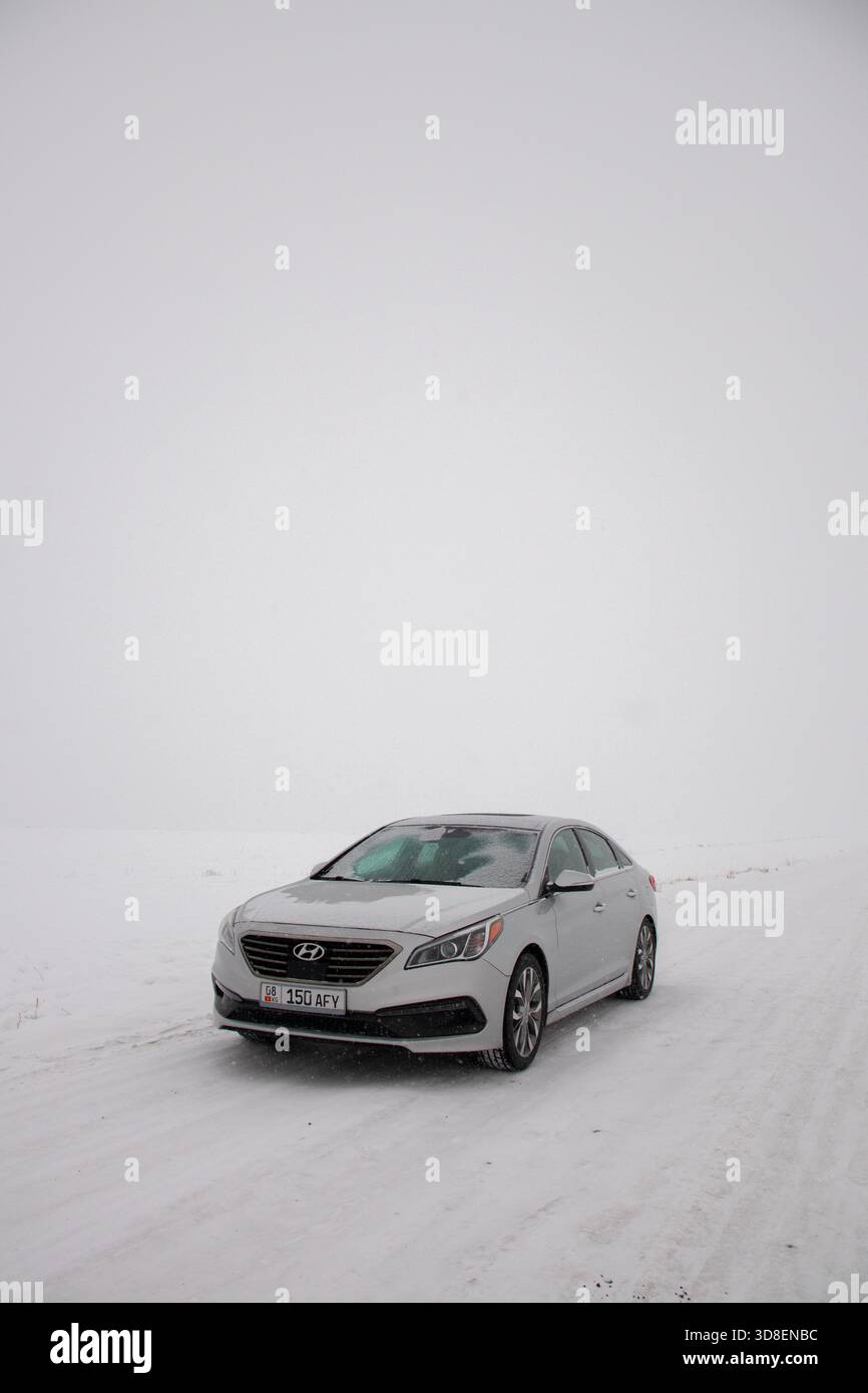 Kirgisistan - 13. Dezember 2024: Vorderansicht einer silbernen Hyundai Sonata Limousine, die auf einer verschneiten Straße unter einem weiten, schroffen weißen Himmel geparkt ist Stockfoto