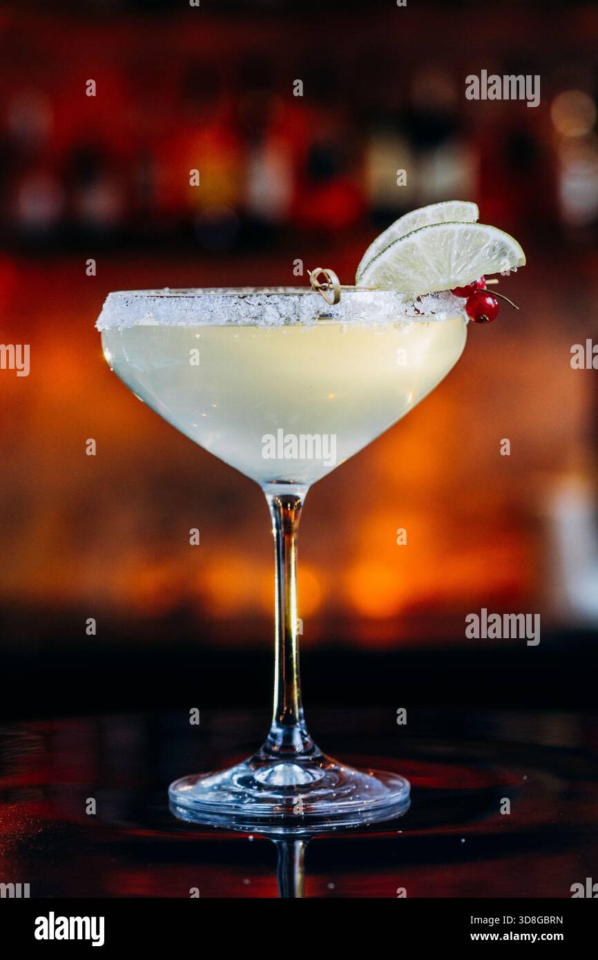 Gelber Cocktail in einem Margarita-Glas mit gesalzenem Rand, Limettenkeilen und roten Beeren. Hintergrund des warmen Balkens. Stockfoto