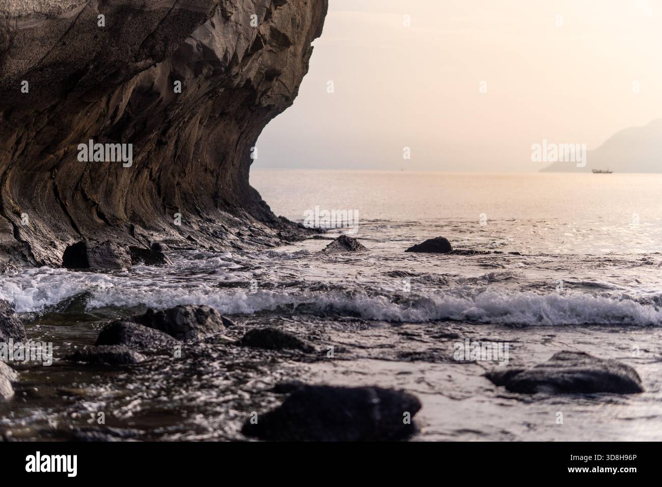 Flacher Blick auf Meereswellen, die in der Nähe einer dramatischen, geschwungenen Klippe auf dunkle Felsen treffen. Ruhige Meereslandschaft mit sanftem Horizontlicht in der Dämmerung. Stockfoto
