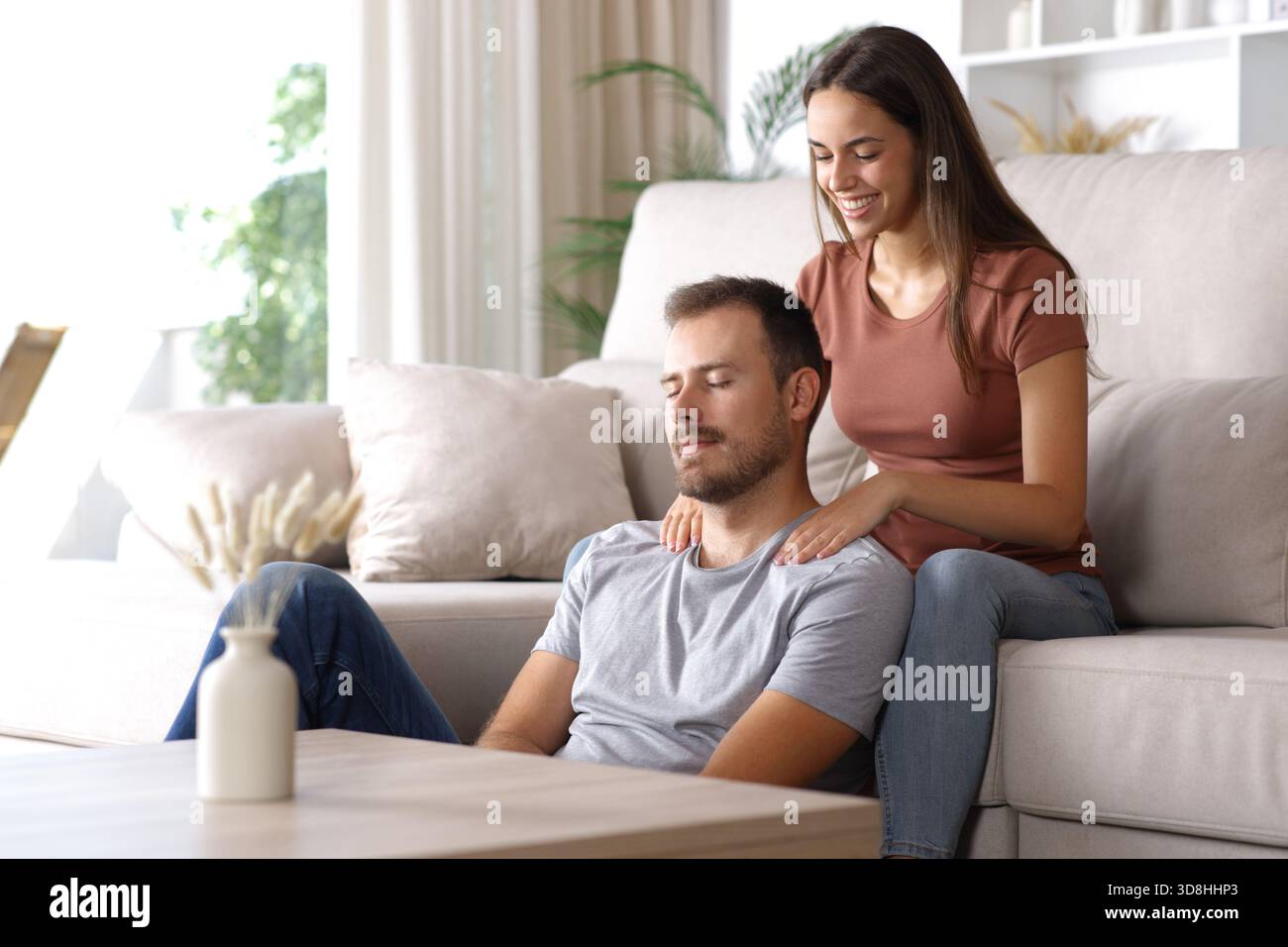 Glückliche Frau massiert ihren entspannten Partner, der zu Hause auf einer Couch sitzt Stockfoto