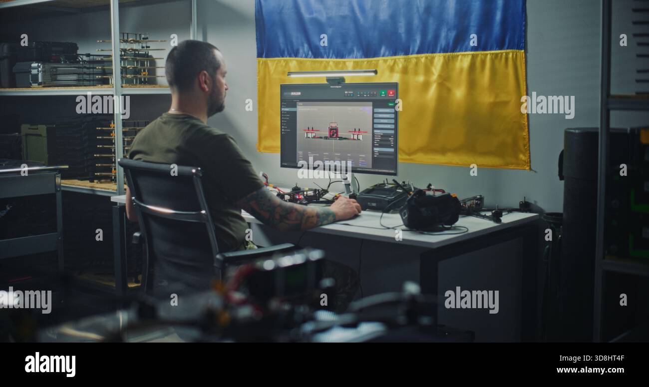 Ukrainischer Ingenieur mit Tattoos überprüft moderne FPV Drohne mit Computer, mit ukrainischer Flagge hängt an der Wand hinter dem Menschen. Konzept der nationalen Bemühungen im Bereich der Verteidigungstechnologien und der Fertigung. Stockfoto