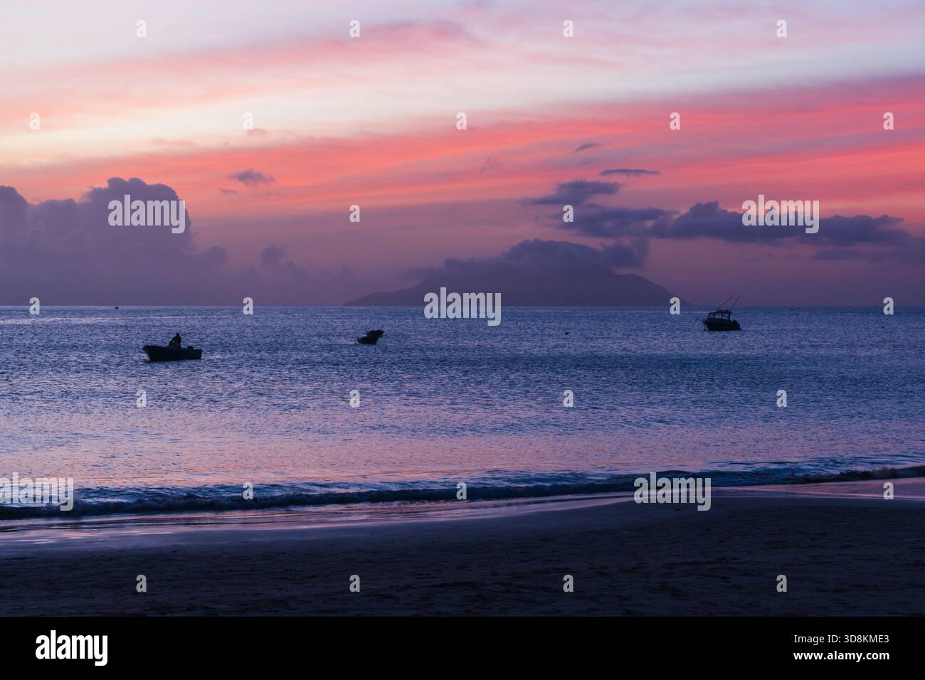 Eine ruhige Küstenszene in der Abenddämmerung mit pastellfarbenen Rosa- und Lila-Tönen, die sich auf sanften Wellen spiegeln. Kleine Boote treiben in der Nähe einer fernen Insel und bilden eine Erbse Stockfoto