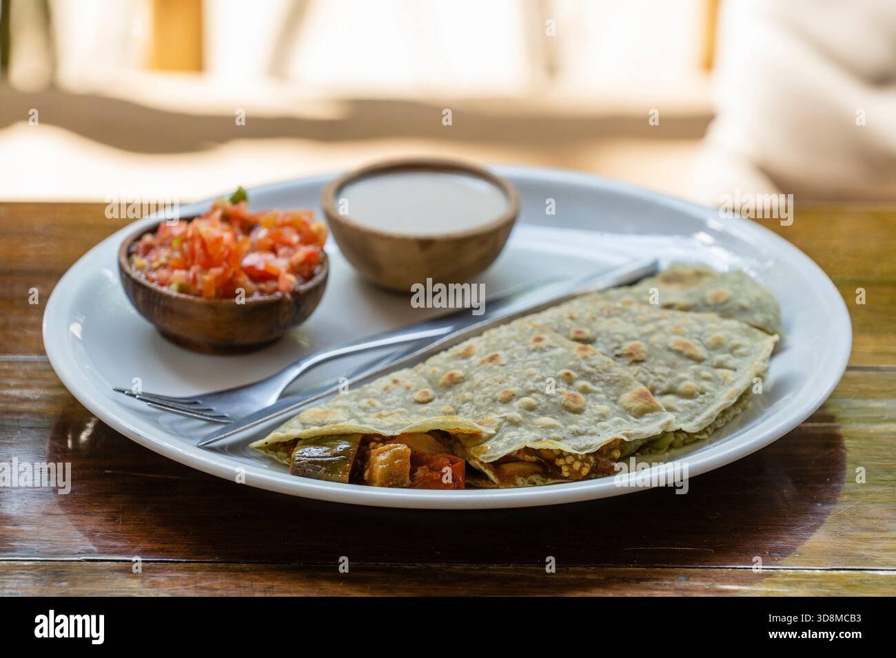 Quesadillas aus Spiruline Tortilla mit Cashewkäse, Auberginen, Tomaten und Sauerrahm und Tomatensalsa auf einem Holztisch im tropischen Café in Thailand, Stockfoto