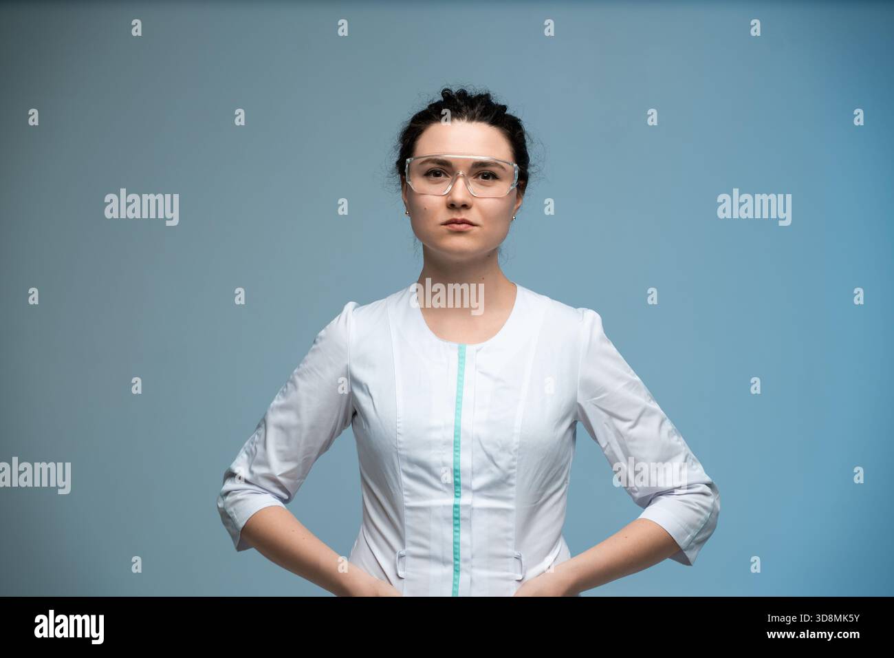 Ärztin in Schutzbrille posiert selbstbewusst mit Händen auf Hüften auf blauem Hintergrund Stockfoto