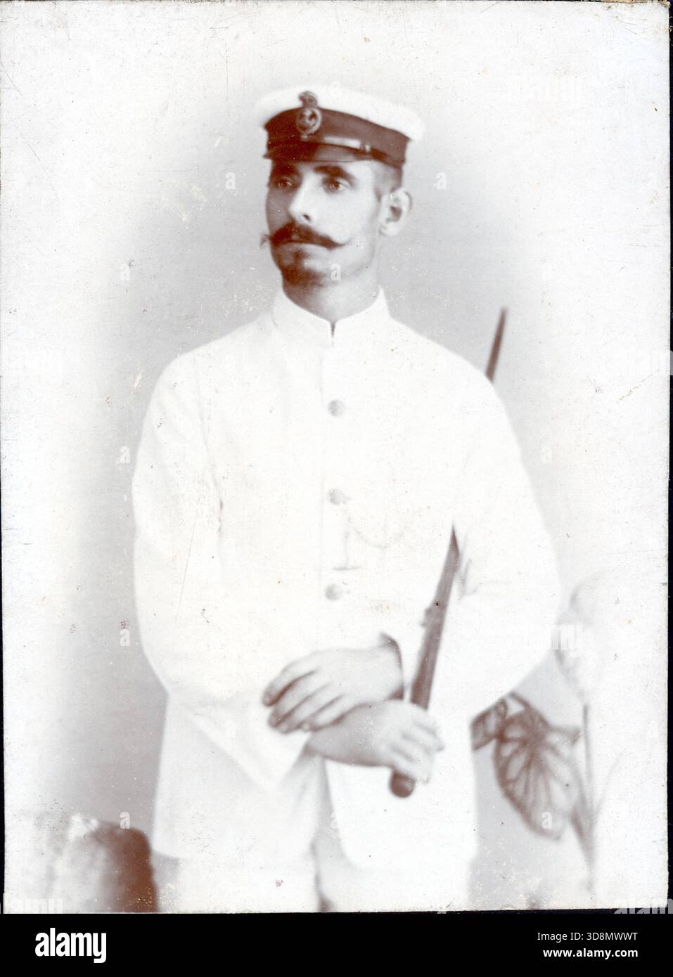 Sepia-Porträt des Mannes in formeller weißer Militäruniform mit Hochkragen und Emblem. Hält einen zeremoniellen Schlagstock mit der rechten Hand. Der schlichte Hintergrund mit schwachen Vorhängen lässt auf spätviktorianische oder edwardische Marinepräsentationen schließen. Von einem späten viktorianischen bis frühen edwardianischen Original-Fotoalbum, das viele Albumenkarten und Visitenkarten enthielt und einen Einblick in die Stile und Moden der Zeit bot. Stockfoto