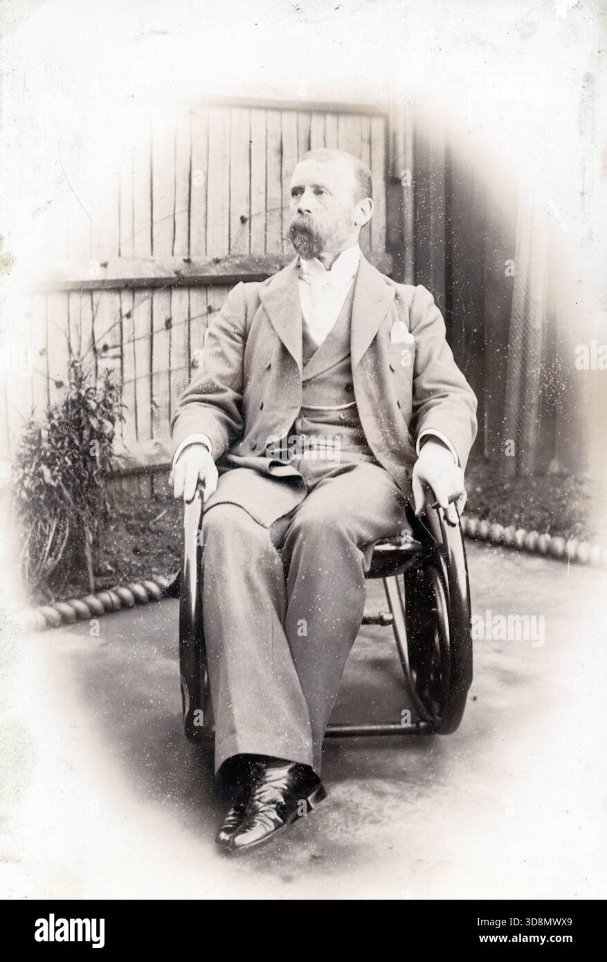Ein bärtiger Mann in einem formellen Anzug sitzt draußen in einem frühen Rollstuhl, eingerahmt von einem Holzzaun und Laub. Die verblasste Vignette und das antike Design deuten auf ein fotografisches Porträt des späten 19. Oder frühen 20. Jahrhunderts hin. Von einem späten viktorianischen bis frühen edwardianischen Original-Fotoalbum, das viele Albumenkarten und Visitenkarten enthielt und einen Einblick in die Stile und Moden der Zeit bot. Stockfoto