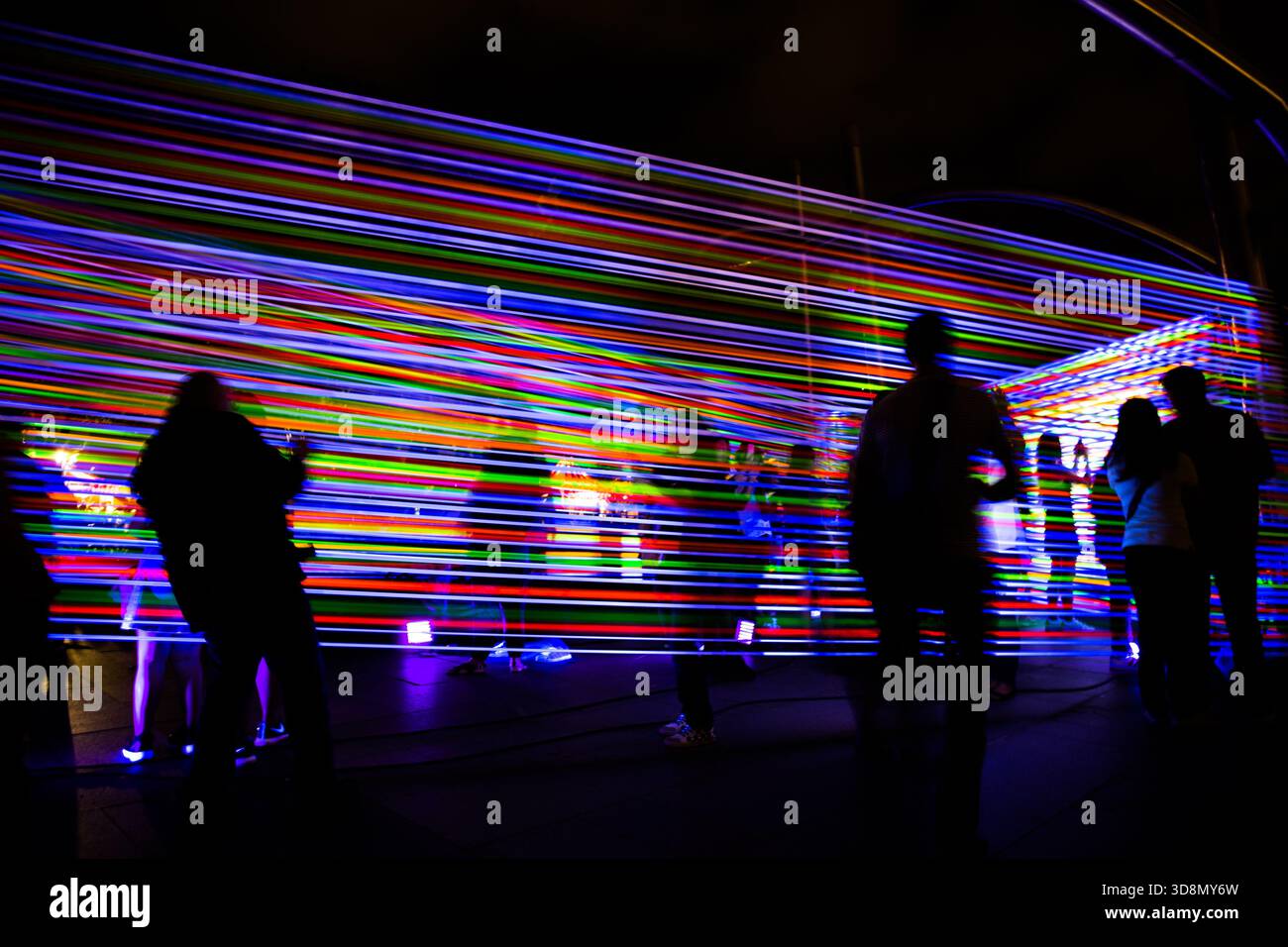 Silhouettenfiguren stehen nachts vor einer blendenden, bunten Lichtwand. Stockfoto