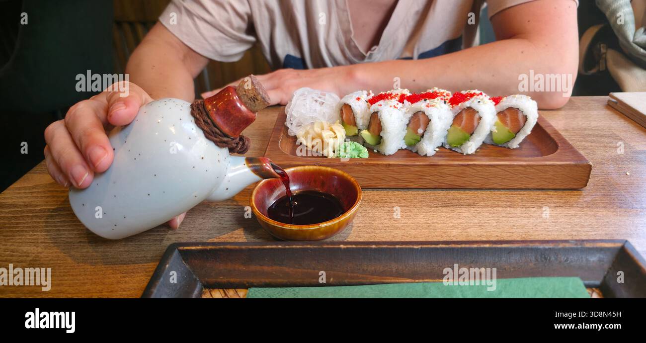 Hand gießt Sojasauce aus einer Keramikflasche auf Sushi-Brötchen, die auf einem Holztisch angeordnet sind, und bietet ein köstliches kulinarisches Erlebnis. - Smartphone-aufgenommenes Stockfoto