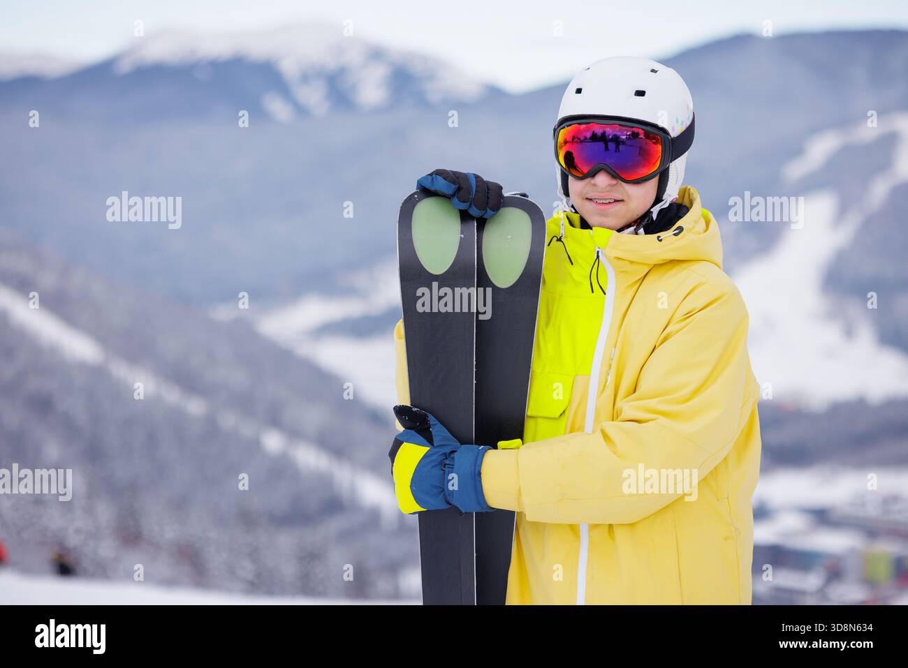 Lächelnder Teenager mit gelbem Skijacken-Helm und Skibrille, die Ski auf Schnee hält, mit verschwommenen Winterbergen dahinter Stockfoto