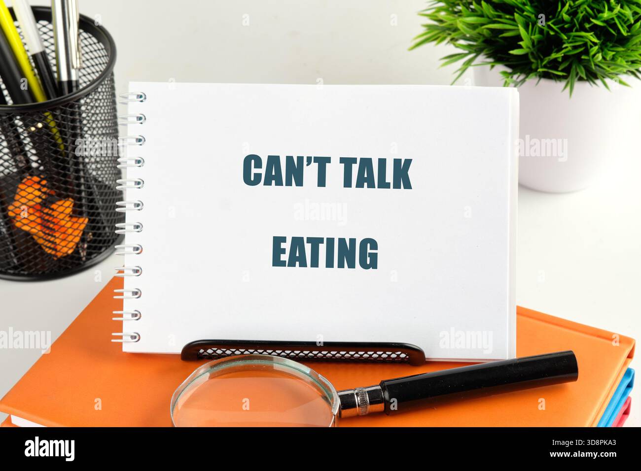 Ein weißes Spiralnotizbuch mit dem Spruch Cant Talk Eating in fetten Buchstaben liegt auf orangefarbenen und blauen Büchern. Ein Stifthalter, eine Pflanze und eine Lupe sind Stockfoto