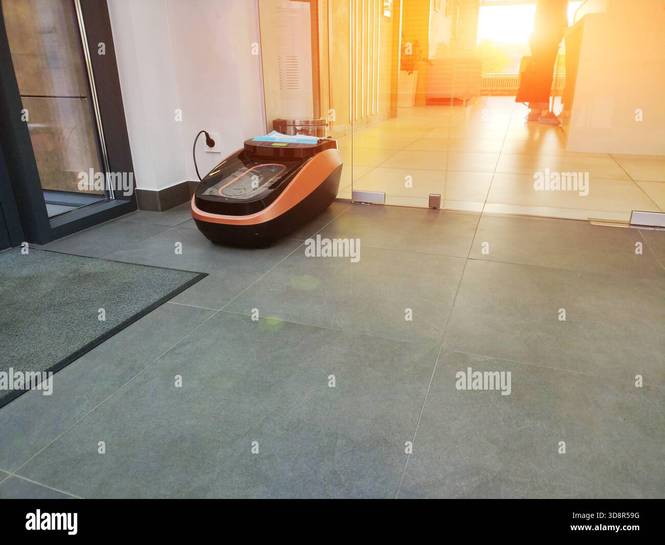 Abroller für Schuhüberzüge auf Fliesenboden im modernen Innenraum, einsatzbereit. Die Person im Hintergrund hat bereits einen blauen Einwegschuh aufgetragen Stockfoto